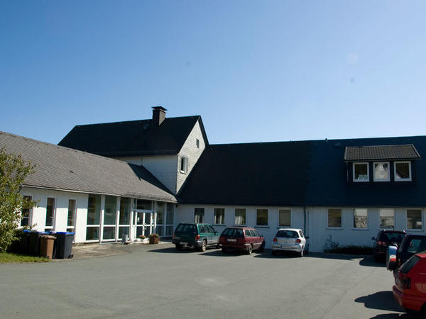 Bild vergr&ouml;&szlig;ern: Blick &uuml;ber den Parkplatz auf das L-f&ouml;rmige Schulgeb&auml;ude der Edertalschule, Verbundschule, Teilstandort Dotzlar. Links im Bild ist der eingeschossige Verwaltungstrakt mit Eingang sichtbar. Die W&auml;nde sind wei&szlig; gestrichen und das Dach ist mit Schiefer eingedeckt. Am linken Ende des Geb&auml;udeteils sind 3 bodentiefe Fenster sichtbar. Vor den Fenstern stehen mehrere M&uuml;lltonnen f&uuml;r Biom&uuml;ll und Altpapier. Nach rechts schauend schlie&szlig;t sich der in einer Glasfront gehaltene Eingangsbereich mit doppelfl&uuml;geliger T&uuml;r an. Die Rahmen sind alle in wei&szlig; gefertigt. Vom Parkplatz aus schaut man &uuml;ber die dort parkenden Autos frontal auf den Klassentrakt. Auch hier ist die Wandfarbe und die Rahmen der Fenster in wei&szlig; gearbeitet. Im Dachgeschoss ist am rechten Bildrand noch eine Schleppgaube mit drei Fenstern zu sehen. Die Gaube ist mit Pfannen eingedeckt. Am rechten Bildrand sind im Vordergrund noch ein paar geparkte Autos erkennbar.