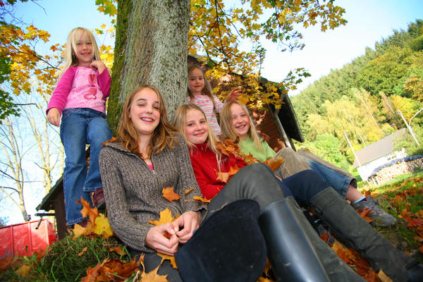 Bild vergrößern: Zu sehen sind mehrere unter einem Baum im Herbstlaub sitzende sowie links und rechts am Stamm stehende Kinder. Bei den Kindern handelt es sich Mädchen verschiedener Altersgruppen welche lächelnd zur Kamera schauen.