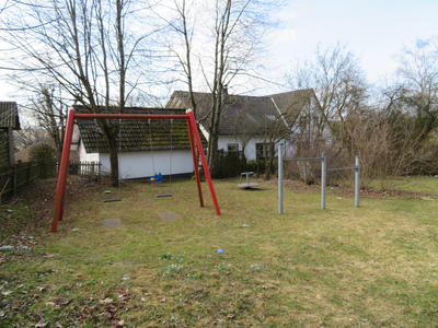 Bild vergr&ouml;&szlig;ern: Das Bild zeigt den Spielplatz in der Ortschaft Rinthe. Im Vordergrund des Bildes ist eine Wiese zu sehen. Schaut man &uuml;ber diese in Richtung des linken Bildrandes sieht man dort eine rote Doppelschaukel welche mit zwei Schaukelbrettern ausgestattet ist. Schaut man von dort etwas nach rechts hin&uuml;ber schaut man auf ein Reck welches mit zwei verschieden hohen Querstangen ausger&uuml;stet ist sehen. In der hinteren rechten Ecke des Spielplatzes steht ein kleines Drehkarussell. An der linken Seite wird der Spielplatz durch einen Zaun gegen das Nachbargrundst&uuml;ck abgegrenzt. Rechts stehen ein paar Str&auml;ucher ohne Laub als Grenze zum Nachbargrundst&uuml;ck. Im Hintergrund wird der Platz durch ein Wohnhaus mit daneben stehenden laublosen B&auml;umen abgeschlossen.