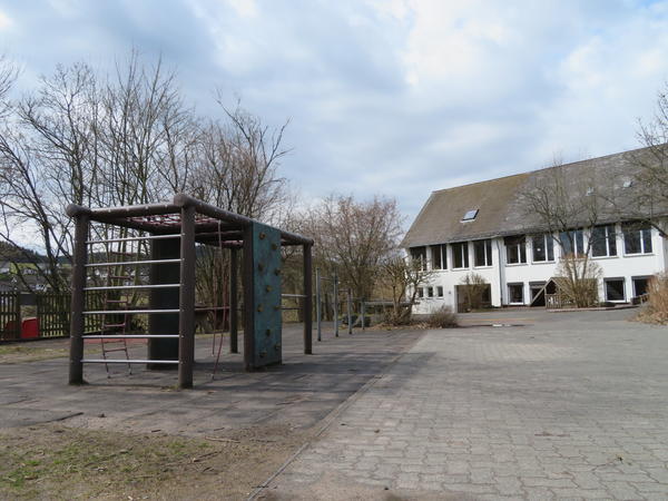 Bild vergr&ouml;&szlig;ern: Man blickt &uuml;ber eine gepflasterte Hoffl&auml;che auf das Schulgeb&auml;ude im Hintergrund. Links im Bild sieht man ein Kletterger&uuml;st auf einer mit Fallschutzmatten ausgelegten Fl&auml;che. Das Kletterger&uuml;st hat an der linken Stirnseite 6 Querstangen und an der linken Frontseite ist eine Leiter aus Tauen angebracht. An der rechten Frontseite ist ein Seil angebracht &uuml;ber diese beiden Seitenteile kann man auf das Ger&uuml;st gelangen welches mit Tauen versehen ist um sich dort hinzusetzen oder hin und her zu laufen. In der Mitte der Vorderseite und der R&uuml;ckseite ist jeweils eine Kletterwand mit Kletterhilfen angebracht. Schaut man an dem Ger&uuml;st vorbei Richtung Geb&auml;ude sieht man auf der Mattenfl&auml;che noch 3 Reckstangen in unterschiedlichen H&ouml;hen stehen.