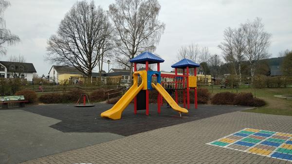 Bild vergr&ouml;&szlig;ern: Zu sehen ist ein Spielplatz auf einer gepflasterten Fl&auml;che. Im Vordergrund ist am rechten Bildrand ein Feld mit H&uuml;pfk&auml;stchen aufgezeichnet. Die einzelnen Felder darin sind in den Farben gr&uuml;n, gelb, braun und blau ausgemalt. Der Rahmen ist in wei&szlig; ausgef&uuml;hrt. Auf der sich anschlie&szlig;enden Fl&auml;che die mit Fallschutzmatten ausgelegt ist steht ein Spielger&auml;t mit 2 T&uuml;rmen. Am linken Turm sind 2 gelbe Rutschen zu sehen. Die beiden T&uuml;rme sind durch ein Kletternetz verbunden. Zur Absturzsicherung sind die Seiten durch ein Netz abgesichert. Hinter dem Turm steht auf der Fl&auml;che noch ein kleines Karussell und ganz am Ende der Fl&auml;che sieht man am linken Bildrand eine Sitzbank aus Holz. Neben der gepflasterten Fl&auml;che schaut man rechts auf eine Wiese, welche durch ein paar Str&auml;ucher abgetrennt wird.