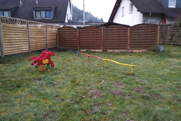 Bild vergr&ouml;&szlig;ern: Man schaut auf eine Rasenfl&auml;che welche von einem braunen Holz-Sichtschutzzaun umfasst ist. Auf der Rasenfl&auml;che ist links ein rotes Wipp-Tier auf einer Feder zu sehen. Etwas davor schaut man auf eine aus Holz gefertigte Strecke zum Balancieren. Die Strecke verl&auml;uft etwas geschwungen und ist in den Farben Gelb, Rot und Blau gefertigt.