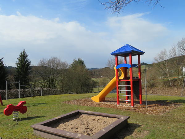 Bild vergr&ouml;&szlig;ern: Man schaut auf den Spielplatz in Ortschaft Schwarzenau der auf einer Wiese angelegt wurde. Mittig des Bildes ist im Vordergrund ein braunen Holzsandkasten zu sehen. Links daneben ist eine rote Feder-Wippe in Form eines Motorrades zu sehen. Hinter dem Sandkasten ist auf einer mit Mulch ausgelegten Fl&auml;che ein Spieleturm zu sehen. Das Gestell ist rot und das Spitzdach ist blau. Als Aufgang ist auf der Frontseite eine Leiter aus vier Querstangen angebracht. Auf der R&uuml;ckseite verf&uuml;gt der Turm &uuml;ber eine Kletterwand. Links am Turm ist eine gelbe Rutsche installiert. Am linke Bildrand ist ein Reck mit drei verschieden hohen Stangen zum Turnen aufgebaut. Dahinter ist ein Holzzaun als Abschluss des Gel&auml;ndes zu sehen. Hinter dem Zaun sind hohe B&auml;ume zu sehen.  Am rechten Bildrand, hinter dem Spieleturm, sieht man auf eine mit B&auml;umen bewachsene B&ouml;schung. Der blaue, mit Schleierwolken verhangene Himmel bildet einen sch&ouml;nen Kontrast zu dem farbintensiven Spielturm.