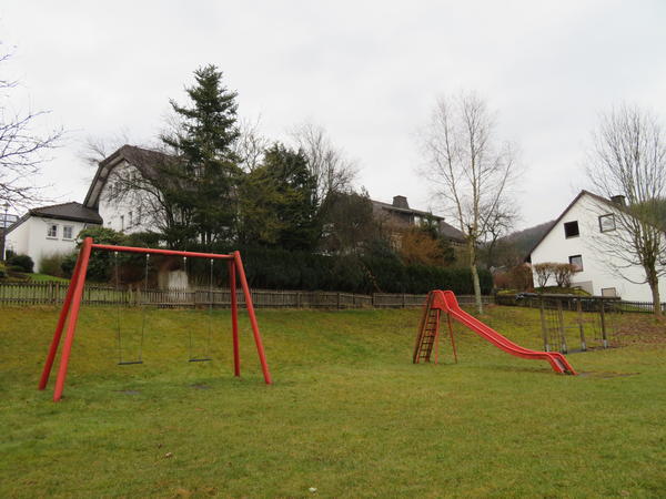Bild vergr&ouml;&szlig;ern: Man schaut &uuml;ber die Wiese auf die vor einer B&ouml;schung stehenden Spielger&auml;te. Hier sind nebeneinander eine rote Doppelschaukel mit zwei Schaukelbrettern, eine rote Rutsche und ein Kletterger&uuml;st zu sehen. Das Kletterger&uuml;st verf&uuml;gt &uuml;ber ein Netz sowie mehreren Tauen zum Klettern. Das Gel&auml;nde wird  auf der B&ouml;schung durch einen Holzzaun abgegrenzt. Hinter dem Zaun sind eine gr&uuml;ne Hecke sowie ein paar hohe Nadelb&auml;ume vor einem Wohnhaus zu sehen. Am rechten Bildrand wird der Spielplatz ebenfalls von einem Holzzaun gegen das anschlie&szlig;ende Grundst&uuml;ck abgegrenzt. In der rechten Spielplatzecke steht eine unbelaubte Birke. Hinter dem Zaun an der linken Bildseite schaut man auf die Giebelseite eines wei&szlig; gestrichenen Wohnhauses.