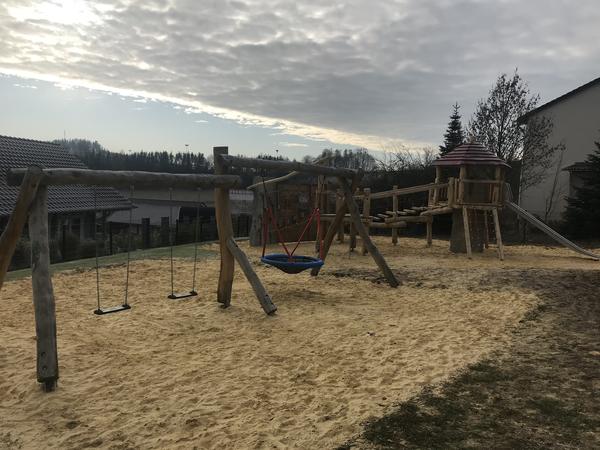 Bild vergr&ouml;&szlig;ern: Hier schaut man auf eine mit Sand ausgelegte Fl&auml;che. Darauf befindet sich links im Vordergrund ein Spielger&auml;t bestehend aus Doppelschaukel mit zwei Schaukelbrettern und einer Nestschaukel. Das Nest ist schwarz mit einer blauen Einfassung. Es ist mit starken roten Seilen befestigt. Im Anschluss daran ist ein Spieleturm zu sehen. Rechts schlie&szlig;t der Turm mit einer Metallrutsche ab. Vorne am Turm befindet sich ein Netz zum Klettern. Von dem Turm kann man nach links &uuml;ber eine Br&uuml;cke das Ger&auml;t verlassen. Die einzelnen Stege der Br&uuml;cke werden von Seilen gehalten. Sie wird durch ein Holzgel&auml;nder abgesichert. Alle Spielger&auml;te sind aus Holz gefertigt. Unterhalb des Spielplatzes ist ein mit Dachpfannen bedecktes Wohnhauses zu sehen. Am rechten Bildrand sieht man hinter dem Spielplatzgel&auml;nde einen Teil eines wei&szlig; gestrichenen Hausgiebels. Zwischen dem Spielplatz und dem Haus stehen ein paar B&auml;ume.