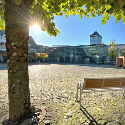 Bild vergrößern: Auf dem Foto ist das Bürgerhaus am Markt in Bad Berleburg in herbstlicher Atmosphäre abgebildet. Im Vordergrund steht eine Bank unter einem Baum, dessen noch grüne Blätter von der Sonne angestrahlt werden. Der Himmel ist wolkenlos.