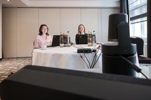 Bild vergrößern: Jessica Durstewitz und Rebecca Dienst sitzen an einem Tisch an ihren Laptops. Per Video sind ihnen die Partner aus Tansania zugeschaltet.