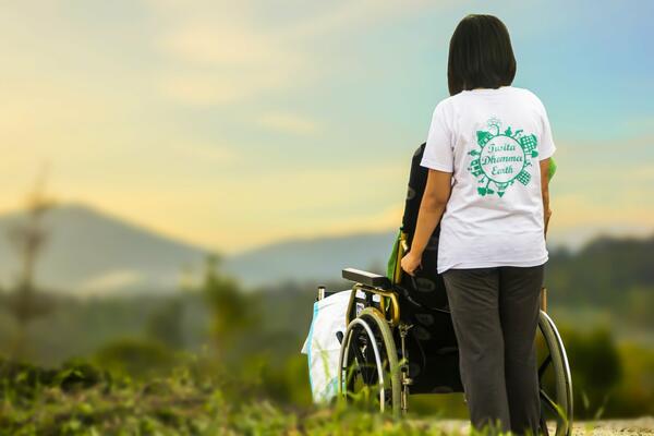 Bild vergrößern: Eine Pflegekraft schiebt eine zu pflegende Person in einem Rollstuhl mit Blick in die Natur.
