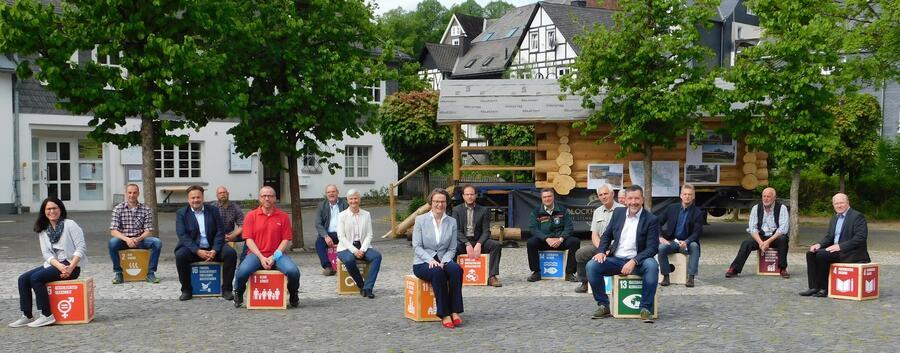 Bild vergrößern: Das Akteursnetzwerk Nachhaltiges Bauen mit Holz sitzt gemeinsam mit der Ministerin für Heimat, Kommunales, Bauen und Gleichstellung Ina Scharrenbach und Bürgermeister Bernd Fuhrmann auf Sitzwürfeln aus Käferholz, auf die die 17 SDG's aufgedruckt sind.