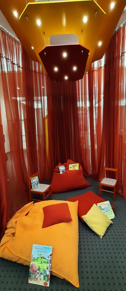 Bild vergr&ouml;&szlig;ern: In der Stadtb&uuml;cherei liegen kleine und gro&szlig;e farbige Kissen auf dem Boden. Auf ihnen liegen B&uuml;cher mit Abenteuern mit Anouk.