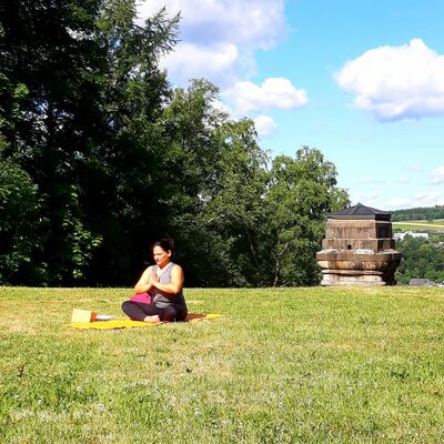 Bild vergrößern: Die Yoga Lehrerin Ale sitzt auf Ihrer Matte und hält die Hände in Gebetsposition vor dem Brustkorb geschlossen. Im Hintergrund blickt man auf die Spitze der Bismarcksäule und die Oberstadt Bad Berleburg.