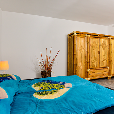 Bild vergrößern: Im Bild sieht man im Vordergrund ein Doppelbett mit einer hellblauen Schlafdecke und im Hintergrund einen hellbraunen Holzschrank.