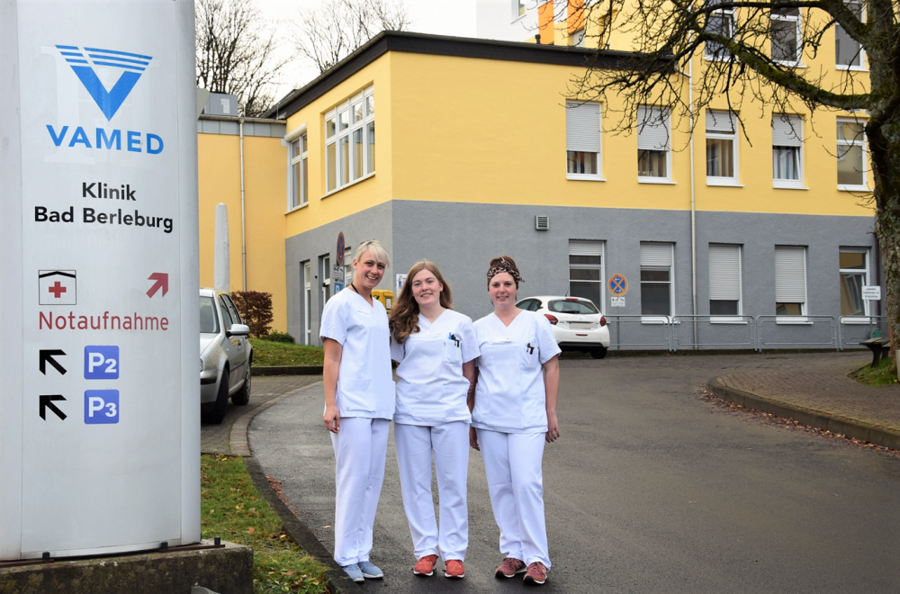 Bild vergrößern: Im Bild sieht man drei junge Frauen, die vor dem Gebäude/Haupteingang der VAMED Akutklinik stehen. Sie sind alle drei in weiß gekleidet.