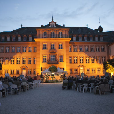 Bild vergrößern: Blick auf das Schloß Berleburg am Abend. Vor dem Schloss sieht man eine beleuchtete Bühne mit Musikern. Links und rechts vor der Bühne sind Sitzreihen mit Besuchern des Konzertes zu sehen.