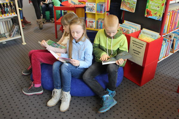Bild vergr&ouml;&szlig;ern: Im Vordergrund sitzen 2 M&auml;dchen und 1 Junge auf einem blauen Sitzsack und lesen. Dahinter ist ein Regal mit B&uuml;chern f&uuml;r Kinder von 6 - 9 Jahren.