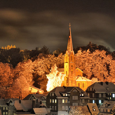 Bild vergrößern: Das Bild zeigt eine Nachtaufnahme der beleuchteten evanglischen Kirche in Bad Berleburg. Vor der Kirche sind Gebäude der zu sehen. Die Kirche selbst wird von Laubbäumen halbkreisförmig umrahmt.