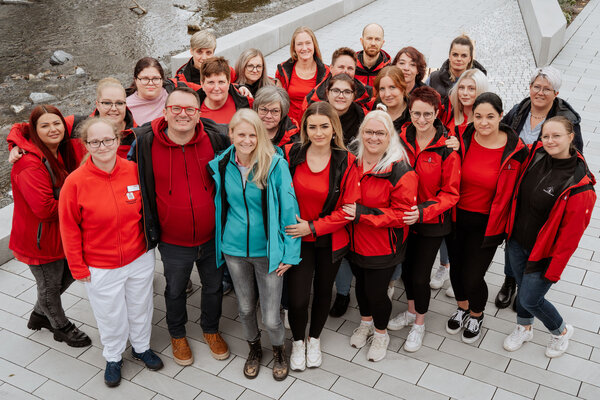 Bild vergr&ouml;&szlig;ern: Im Bild sieht man das gesamte Team der Mobilen Schwestern. Alle Personen sind mit einer roten Jacke mit schwarz abgesetzt bekleidet. Die Leitung steht im Vordergrund und tr&auml;gt eine Jacke in t&uuml;rkis.