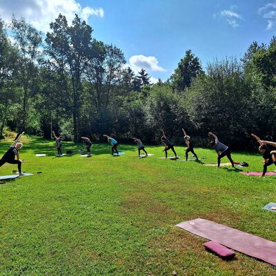 Bild vergr&ouml;&szlig;ern: Im Bild sieht man auf der linken Seite die Yoga Lehrerin und im Halbkreis auf der rechten Seite stehend die Gruppe. Sie sind alle in einer Vorbeuge ausgerichtet und haltet diese Dehnungsposition.