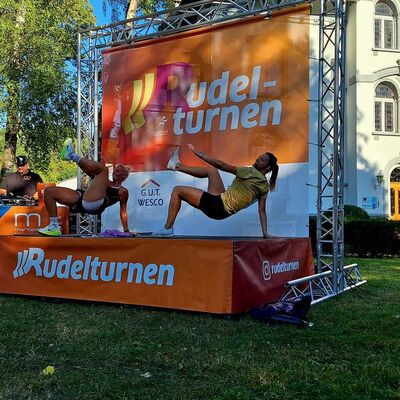 Bild vergr&ouml;&szlig;ern: Im Bild sieht man die zwei Akteure Maria und Sarah auf der B&uuml;hne. Sie zeigen gerade eine &Uuml;bung im St&uuml;tz. Hinter der B&uuml;hne sieht man die Jugendstilvilla im Rathauspark.