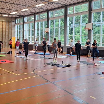 Bild vergr&ouml;&szlig;ern: Im Bild sieht man eine gro&szlig;e Gruppe an Sportlerinnen und Sportlern in einer Turnhalle. Sie sind alle nach vorne ausgerichtet und bewegen sich gerade mit angewinkelten Beinen im Wechsel auf der Stelle.
