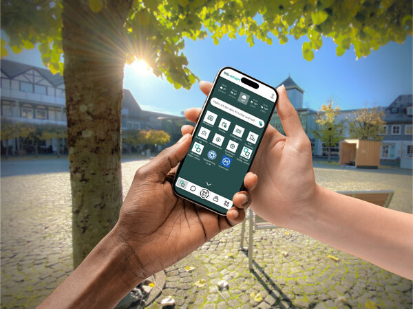 Zwei Personen mit unterschiedlicher Hautfarbe halten gemeinsam ein Smartphone. Auf dem Bildschirm ist die Startseite der App "blbsmart" mit verschiedenen digitalen B&uuml;rgerservices zu sehen. Im Hintergrund liegt ein sonniger, gepflasterter Marktplatz mit Fachwerkh&auml;usern und gr&uuml;nen B&auml;umen.