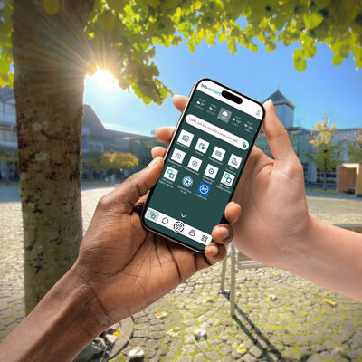 Zwei Personen mit unterschiedlicher Hautfarbe halten gemeinsam ein Smartphone. Auf dem Bildschirm ist die Startseite der App "blbsmart" mit verschiedenen digitalen B&uuml;rgerservices zu sehen. Im Hintergrund liegt ein sonniger, gepflasterter Marktplatz mit Fachwerkh&auml;usern und gr&uuml;nen B&auml;umen.