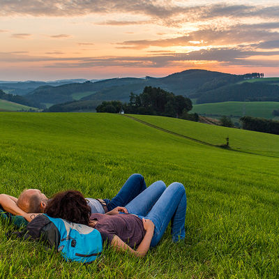 Bild vergrößern: Zwei Wanderer machen eine Rast auf der Wiese und genießen den Sonnenuntergang.