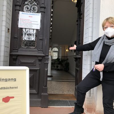 Bild vergr&ouml;&szlig;ern: Die Leiterin der Stadtb&uuml;cherei von Bad Berleburg, Gabriele Klotz, steht vor einer gro&szlig;en Holzt&uuml;r und zeigt mit ihrer Hand auf den neuen Eingang zur Stadtb&uuml;cherei. Dieser f&uuml;hrt durch die gro&szlig;e Holzt&uuml;r. Direkt vor der Holzt&uuml;r steht ein Hinweisschild mit der Aufschrift "Eingang Stadtb&uuml;cherei" sowie einer aufgedruckten roten Hand, die mit dem Zeigefinger auf den Eingang zur Stadtb&uuml;cherei zeigt.