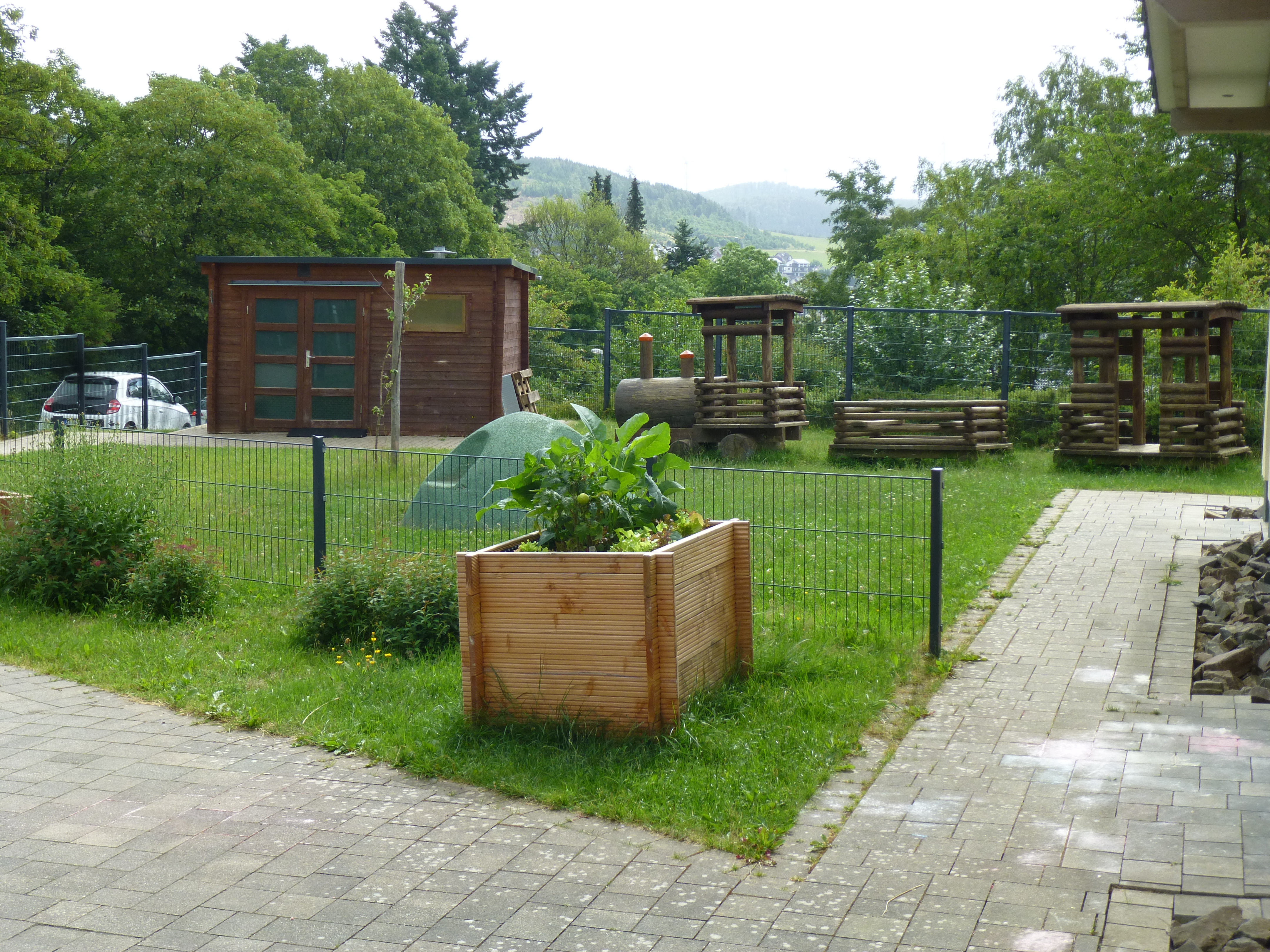 Man sieht den Spielplatz der KIndertageseinrichtung Senfkorn. Auf der gr&uuml;nen Wiese steht links ein Holzschuppen und rechts daneben Holzspielger&auml;te in Form einer Lokomotive. Die Grasfl&auml;che ist umrahmt mit einem Gitterzaun. Vor dem Zaun steht einer gro&szlig;er Holzk&uuml;bel mit Pflanzen. 


&uuml;ber eine gepflasterte Fl&auml;che direkt auf den etwas zur&uuml;ckversetzten Eingang der Kindertagesst&auml;tte. Die Hauswand ist einem Gelbton gestrichen. Der Abschluss des Anstriches unten ist in Grau gew&auml;hlt. Links im Bild sieht man an der Hauswand ein rundes Schild mit dem Logo und dem Schriftzug Evangelische Kindertagesst&auml;tte Senfkorn. Im oberen Bereich ist das Schild wei&szlig;, das Logo sowie der Schriftzug sind in einem Gr&uuml;nton gehalten. Entlang der Hauswand hat man eine Reihe mit kleinen Steinen angebracht. Bei der &Uuml;berdachung des Eingangs sind braune Holzbalken zu erkennen. Die T&uuml;r besteht aus drei Fl&uuml;geln. Der mittlere Fl&uuml;gel ist die T&uuml;r selbst. Oberhalb der Teile sieht man Oberlichter, welche der Breite der Fl&uuml;gel entsprechen. Alle Teile sind vollst&auml;ndig aus Glas und lediglich die Rahmen sind wei&szlig;. Als T&uuml;rgriff ist eine von unten bis oben durchgehende Stange angebracht. Vor dem rechten Seitenteil stehen zwei mit Stiefm&uuml;tterchen bepflanzte quadratische Blumenk&uuml;bel 