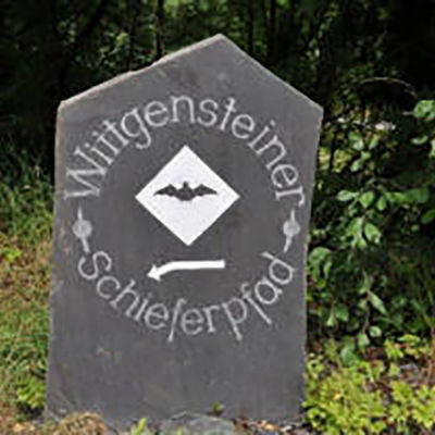 Bild vergrößern: Auf einer Schieferplatte eist der Zuweg zum Wittgensteiner Schieferpfad beschildert. Die Platte zeigt das Wanderwegsymbol, eine Fledermaus auf weißem Grund.