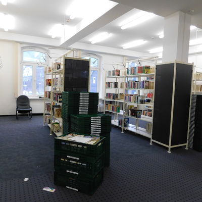 Bild vergrößern: In einem Raum stehen zwei Regale. In den Regalen stehen Bücher. Vor dne Regalen stehen viele grüne Körbe. Zwei Körbe sind mit Büchern gefüllt.