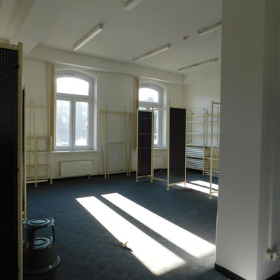 Bild vergrößern: Ein Raum mit zwei Fenstern. Die Fenster werfen Sonnenlicht in den Raum. Im Raum stehen vier Regale. Die Regale sind leer.