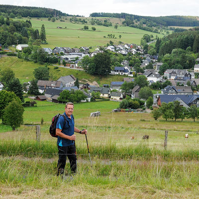 Bild vergrößern: Ein Wanderer mit Rucksack und Wanderstock posiert vor der Ortschaft Wunderthausen